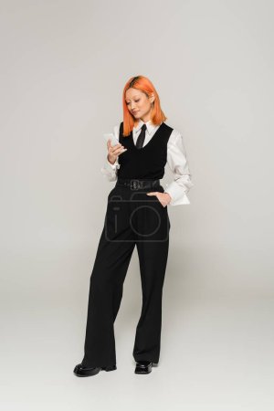 full length of trendy asian woman with dyed red hair standing with hand in pocket and networking on mobile phone on grey background, business casual fashion, white shirt, black vest and pants