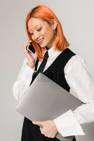 positive emotion, young asian woman with radiant smile and colored red hair, in business casual clothes holding laptop and talking on smartphone on grey background, freelance lifestyle