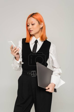 freelance lifestyle, business casual fashion, red haired asian woman in white shirt, black tie and vest standing with laptop and looking at mobile phone on grey background, generation z