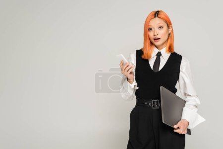 étonnement, surpris femme asiatique avec les cheveux rouges teints et bouche ouverte debout avec ordinateur portable et smartphone tout en regardant la caméra sur fond gris, chemise blanche, gilet noir et pantalon, affaires casual