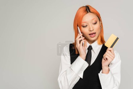 encantadora y elegante mujer asiática con pelo rojo celebración de la tarjeta de crédito y hablando en el teléfono móvil sobre fondo gris, camisa blanca, chaleco negro y corbata, estilo casual de negocios, generación z