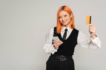 positive emotion, joyful asian woman with dyed red hair holding smartphone, looking at camera and showing credit card on grey background, business casual, white shirt, black tie and vest, gen z