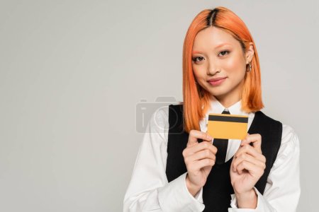 attractive and positive asian woman with dyed red hair smiling and showing credit card on grey background, white shirt, black vest and tie, business casual fashion, generation z