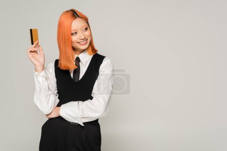 Foto de Feliz emoción, sonrisa alegre, mujer asiática pelirroja sosteniendo tarjeta de crédito y mirando hacia otro lado sobre fondo gris, estilo casual de negocios, camisa blanca, corbata y chaleco negro, generación z - Imagen libre de derechos