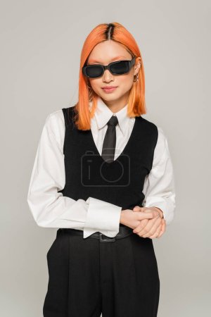 youthful and fashionable asian woman with colored red hair posing in dark sunglasses, white shirt, black tie and vest on grey background, business casual style, generation z