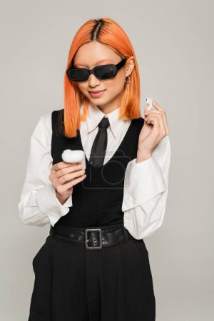 Foto de Mujer asiática sonriente con el pelo rojo teñido mirando el caso con auriculares inalámbricos sobre fondo gris, emoción positiva, gafas de sol oscuras, camisa blanca, corbata y chaleco negro, moda casual de negocios - Imagen libre de derechos