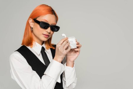 modern music lover, expressive asian woman with dyed red hair, in dark sunglasses, white shirt, black tie and vest showing case with wireless earphones on grey background, gen z lifestyle
