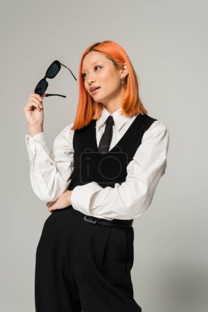 happy face, positive emotion, youthful asian woman with colored red hair holding dark sunglasses and looking away on grey background, black and white clothes, business casual style