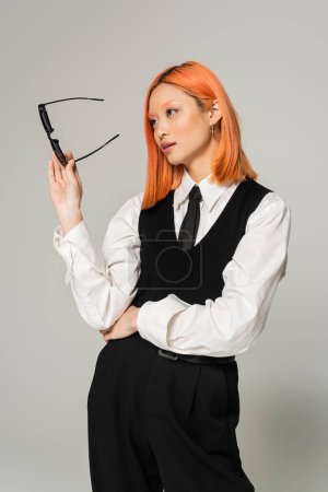 business casual fashion, attractive asian woman with colored red hair, in white shirt, black pants, vest and tie holding dark sunglasses and looking away on grey background, generation z