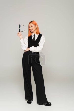 Photo for Happy emotion, full length of asian woman with dyed red hair holding dark sunglasses and looking away on grey background, business casual fashion, white shirt, black tie, vest and pants - Royalty Free Image