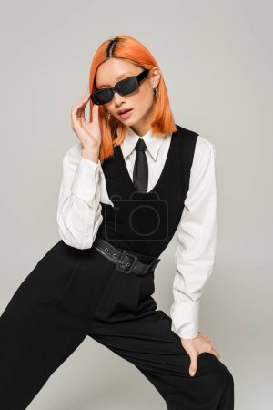fashionable and young asian woman in white shirt, black tie, vest and pants standing in expressive pose and adjusting dark sunglasses on grey background, modern lifestyle, generation z