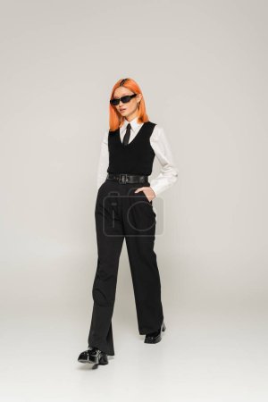 Photo for Full length of stylish asian woman with dyed red hair, in dark sunglasses walking with hand in pocket on grey background, business fashion, white shirt, vest and pants, modern lifestyle - Royalty Free Image