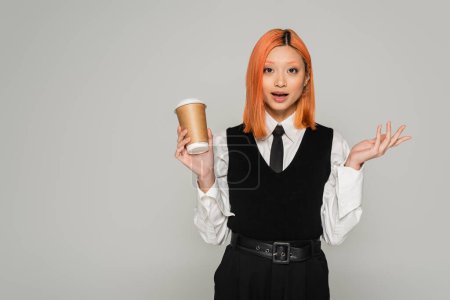 amazement, surprised asian woman with open mouth holding paper cup, gesturing and looking at camera on grey background, business casual style, white shirt, black tie and vest, colored red hair