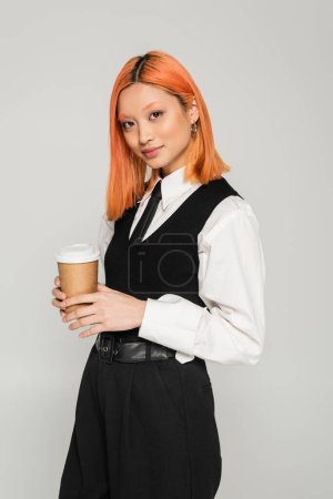 attractive asian woman with colored red hair holding paper cup with takeout drink while looking at camera on grey background, stylish black and white clothes, business casual, gen z