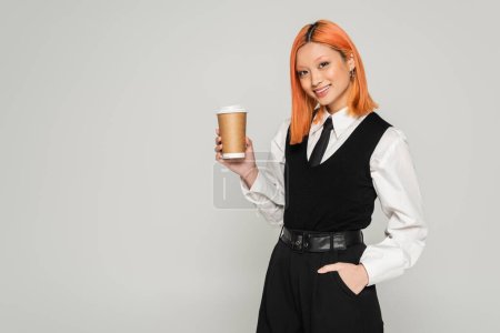 positive emotion, young asian woman with dyed red hair and radiant smile holding hot drink and looking at camera on grey background, black and white business casual clothes, modern fashion