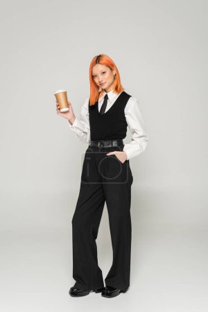 full length of trendy asian woman with dyed red hair smiling at camera while holding takeaway drink on grey background, black vest and pants, white shirt, hand in pocket, business casual style