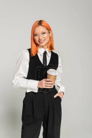 cheerful and young asian woman with colored red hair and trendy eyeglasses standing with hand in pocket and takeaway drink on grey background, business casual, black and white clothes