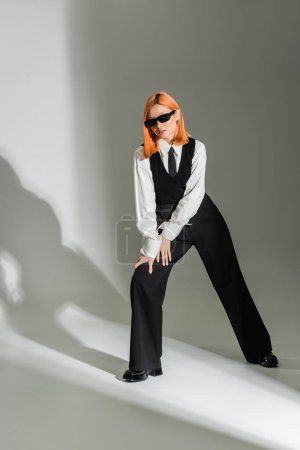 stilvolle Pose einer modischen Asiatin mit gefärbten roten Haaren, dunkler Sonnenbrille, weißem Hemd, schwarzer Krawatte, Weste und Hose auf grauem Hintergrund, Business-Modefotografie, Generation z