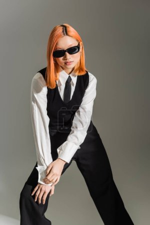 schwarze und weiße Kleidung, dunkle Sonnenbrille, asiatische Frau mit gefärbten roten Haaren, die in stilvoller Pose auf grauem Hintergrund steht, Business-Mode, Generation z Lifestyle