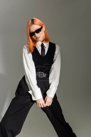 fashionable and youthful asian woman in dark trendy sunglasses standing and posing on grey shaded background, dyed red hair, white shirt, black tie, vest and pants, business casual style