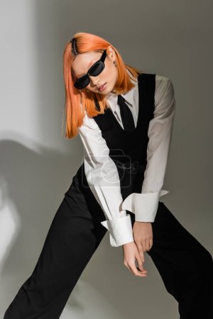 youthful asian fashion model in dark sunglasses standing in stylish pose on grey shaded background, colored red hair, white shirt, black tie, vest and pants, business fashion photography