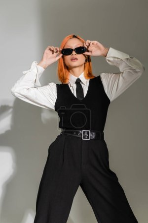 fashion shoot of expressive asian woman with dyed red hair adjusting dark sunglasses while standing in white shirt, black tie, vest and pants on grey shaded background, business casual style