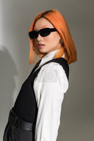 portrait of young and fashionable asian woman in dark sunglasses, white shirt and black vest looking at camera while posing on grey shaded background, generation z lifestyle
