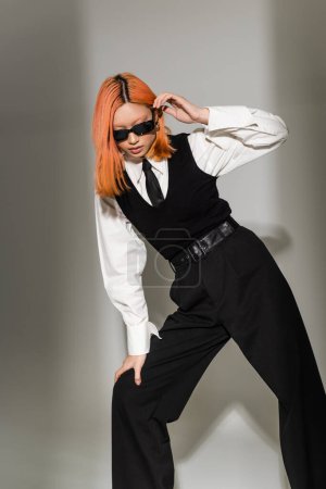 Mode-Shooting moderner asiatischer Frau mit dunkler Sonnenbrille und schwarz-weißer Kleidung auf grauem Hintergrund, gefärbtem roten Haar, weißem Hemd, schwarzer Krawatte, Weste und Hose, stilvoller Pose, Business Casual