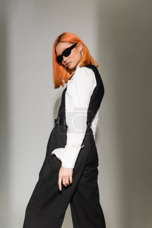 fashion photography, sensual and young asian woman with colored red hair standing and looking at camera on grey shaded background, dark sunglasses, white shirt, black vest, modern fashion shoot