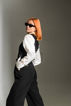 femme asiatique aux cheveux rouges et expressifs en lunettes de soleil sombres, chemise blanche, gilet et pantalon tenant la main dans les poches et regardant la caméra tout en posant sur fond gris ombragé, mode d'affaires moderne