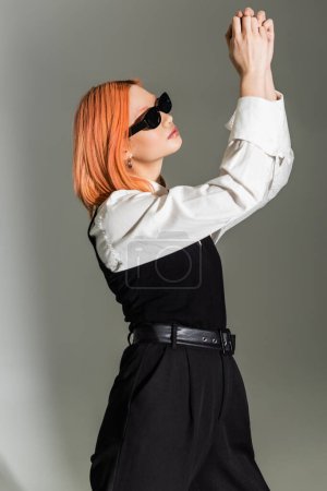 Photo for Side view of expressive and red haired asian woman in dark sunglasses, black and white clothes posing with raised hands on grey shaded background, business casual fashion, generation z - Royalty Free Image
