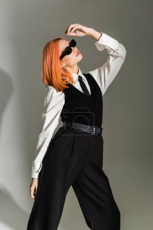 red haired and expressive asian woman with colored hair posing in dark stylish sunglasses, white shirt, black tie, vest and pants on grey shaded background, business casual fashion, generation z