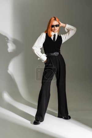 business fashion, trendy asian woman with colored red hair posing with hand in pocket on grey shaded background, dark sunglasses, white shirt, black tie, vest and pants, generation z, full length