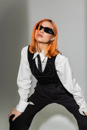 youthful, sensual and attractive asian woman in dark sunglasses, with colored red hair posing in white shirt, black tie, vest and pants on grey shaded background, business fashion photography