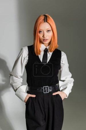 trendy and confident asian woman with colored hair holding hands in pockets and looking at camera on grey shaded background, white shirt, black tie, vest and pants, business casual fashion