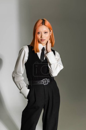 business casual fashion, young asian woman with colored red hair, in white shirt, black vest and pants posing with hand in pocket while looking at camera on grey shaded background, fashion shoot