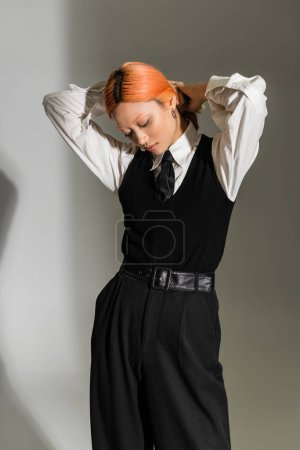 modische asiatische Frau mit gefärbten roten Haaren, in schwarz-weißer Business-Casual-Kleidung posiert mit den Händen hinter dem Kopf auf grauem Hintergrund, jugendlicher Stil, Generation Z, Jugendkultur