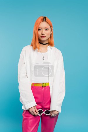 junges asiatisches Model in weißem Hemd, pinkfarbener Hose und trendigen Accessoires mit Sonnenbrille und Blick in die Kamera auf blauem Hintergrund, trendiger Sommer, jugendlicher Stil, Generation z