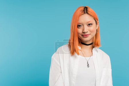 happy emotion, summer vibes, young and charming asian woman with colored red hair looking at camera on blue background, white shirt, necklaces, radiant smile, generation z lifestyle