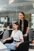 happy hairdresser and female client in beauty salon, cheerful beauty worker with braids standing near tattooed woman, discussing hair treatment, hair extension, customer satisfaction  magic mug #660916336