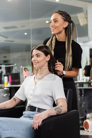 happy hair stylist and female client in beauty salon, beauty worker with braids standing near tattooed woman, discussing hair treatment, hair extension, customer satisfaction, hair care 