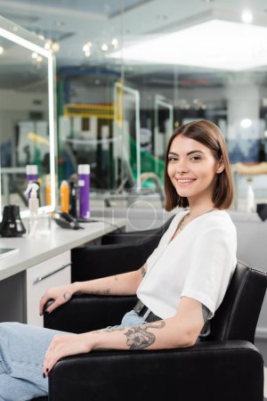 happy female client, tattooed young woman sitting in hairdressing chair and looking at camera in hair salon, beauty service, hair extension, hair coloring, blurred background, generation z 