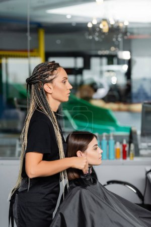 Photo for Salon job, beauty worker wearing hairdressing collar on female client in beauty salon, hairstyling, hairdo, extension, hair treatment, salon customer, beauty profession, side view - Royalty Free Image