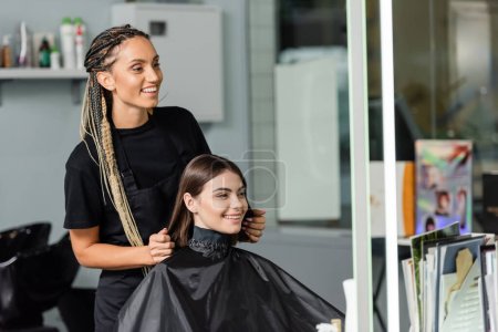Photo for Beauty industry, positivity, happy hair stylist with braids wearing hairdressing cape on female client, hair extension, hair treatment, salon customer, beauty profession - Royalty Free Image