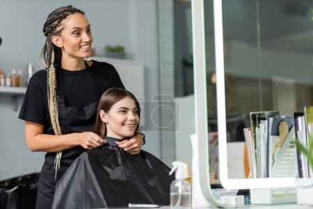 Schönheitsindustrie, Friseur mit Zöpfen Befestigung Friseur Umhang auf weibliche Klientin, Haarverlängerung, Haarbehandlung, Salonkunde, Beauty-Beruf, Salonarbeit, zusammen lächeln 
