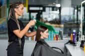 hairstylist spraying hair of female client, hairdresser with braids holding spray bottle near woman with short brunette hair in salon, hair cut, hair treatment, hair make over, hairdo, side view Sweatshirt #660916692