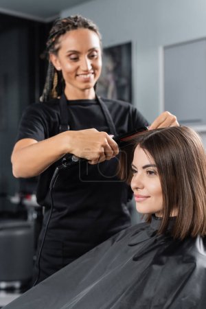 satisfaction du client, femme brune joyeuse avec les cheveux courts dans le salon de beauté, coiffeur professionnel avec lisseur de cheveux coiffant les cheveux de la cliente, travailleur de beauté  