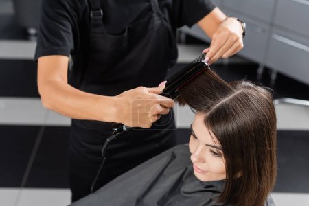 Draufsicht auf professionelle Friseur mit Haarglätter Styling Haare der weiblichen Kunden, Beauty-Arbeiter, Kundenzufriedenheit, brünette Frau mit kurzen Haaren, Schönheitssalon, Haarmode 
