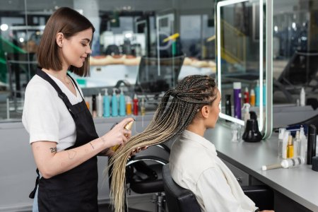 Photo for Hair salon, tattooed beauty worker in apron applying hair product on braids of woman in salon, spray bottle, happy beauty worker, client satisfaction, salon customer, smile, side view - Royalty Free Image
