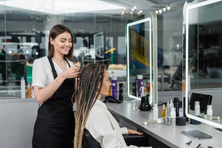 braids, cheerful hairdresser braiding hair of woman in salon, braiding process, salon customer, beauty profession, client satisfaction, hair fashion, hairdo, tattooed, beauty profession 
